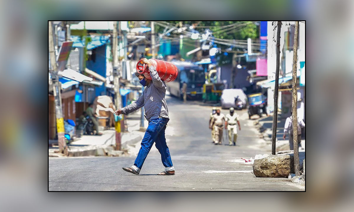 A resident carrying LPG cylinder crosses a road as policemen, seen in the background, march in the area of Padarayanapura in Chamarajapet during ongoing COVID-19 lockdown, in Bengaluru. (PTI)